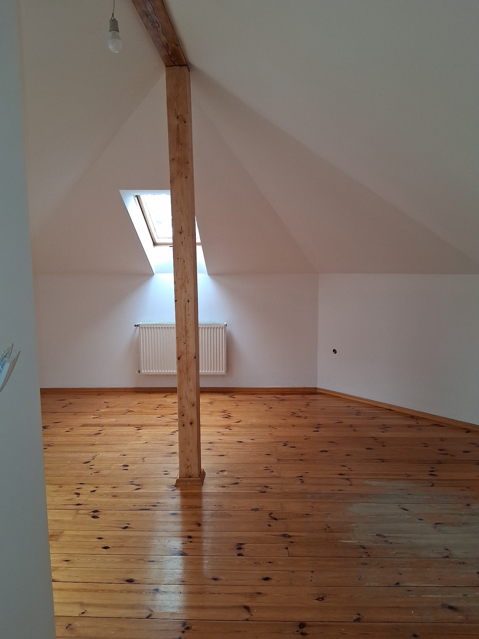 Jasne poddasze z drewnianą podłogą i belką konstrukcyjną. Widoczne okno dachowe i grzejnik. Świeżo pomalowane ściany. W tle widoczna żarówka.