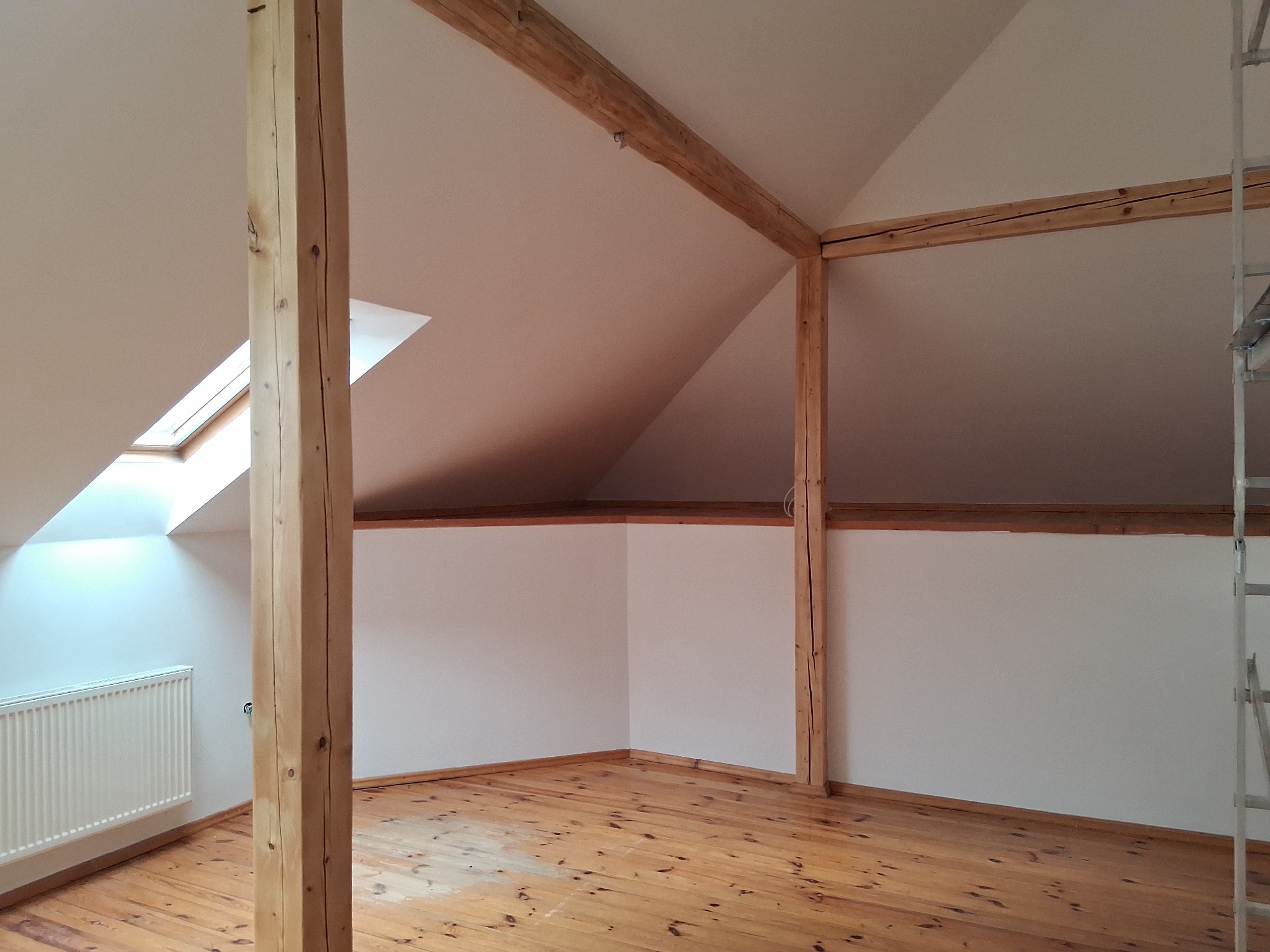 Jasne poddasze po remoncie: drewniana podłoga, białe ściany, drewniane belki stropowe, okno dachowe. Widoczny fragment drabiny malarskiej.