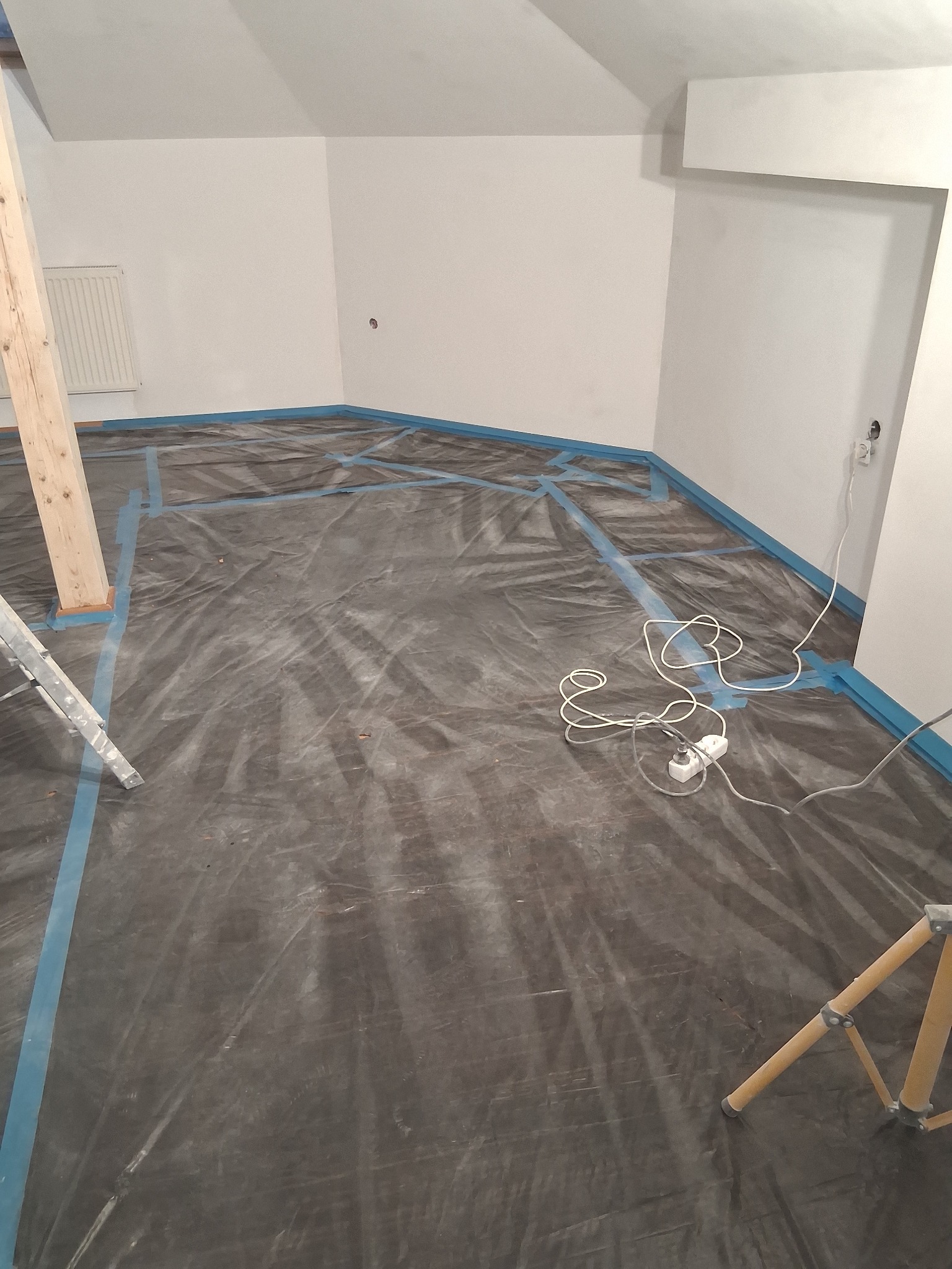 Przygotowanie do malowania: podłoga zabezpieczona folią i taśmą malarską, widoczny fragment drabiny i trójnóg, gniazdko elektryczne z przedłużaczem na podłodze.
