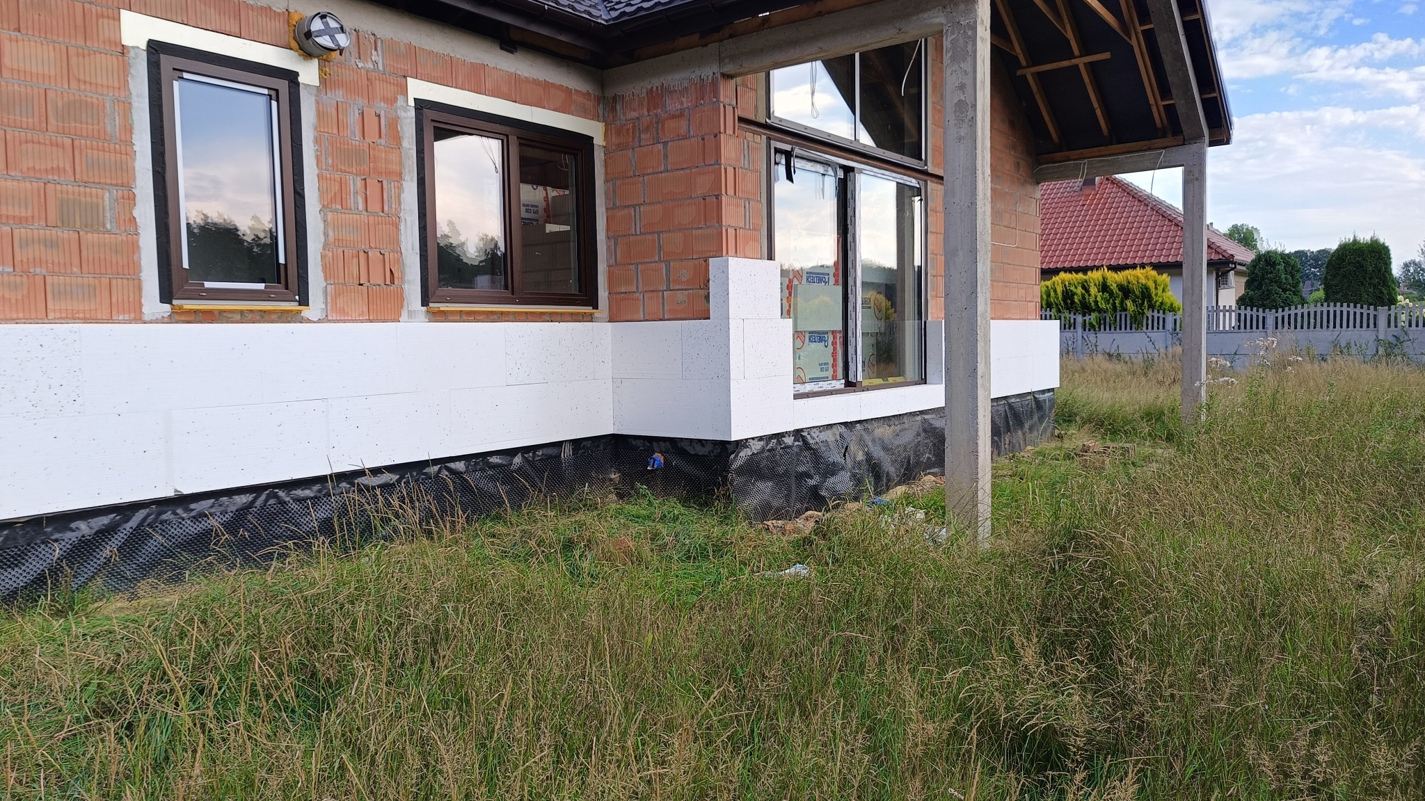 Ocieplenie fundamentów budynku z widoczną folią kubełkową, styropianem i fragmentem elewacji z oknami w trakcie montażu.