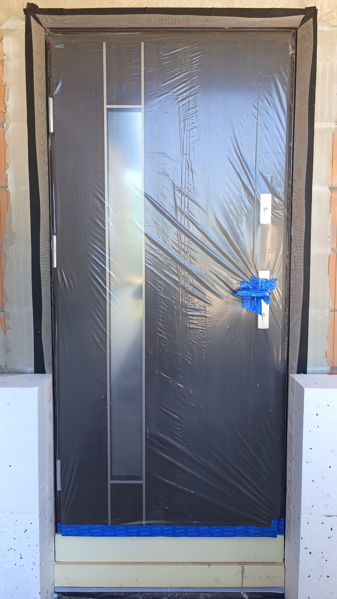 Ciemne drzwi wejściowe z szybą, zabezpieczone folią ochronną i taśmą malarską, zamontowane w surowym otworze ściany z widocznym ociepleniem i siatką elewacyjną.
