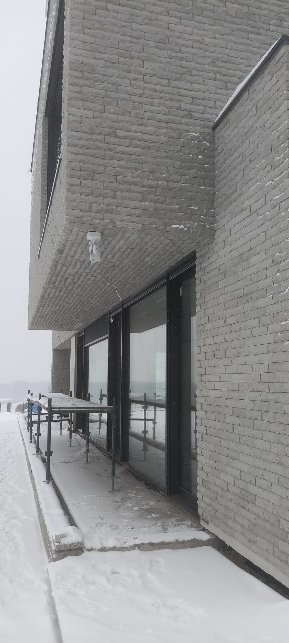 Nowoczesny budynek z elewacją z szarej cegły w zimowej scenerii, częściowo zasłonięty rusztowaniem, z widoczną kamerą zewnętrzną pod balkonem.