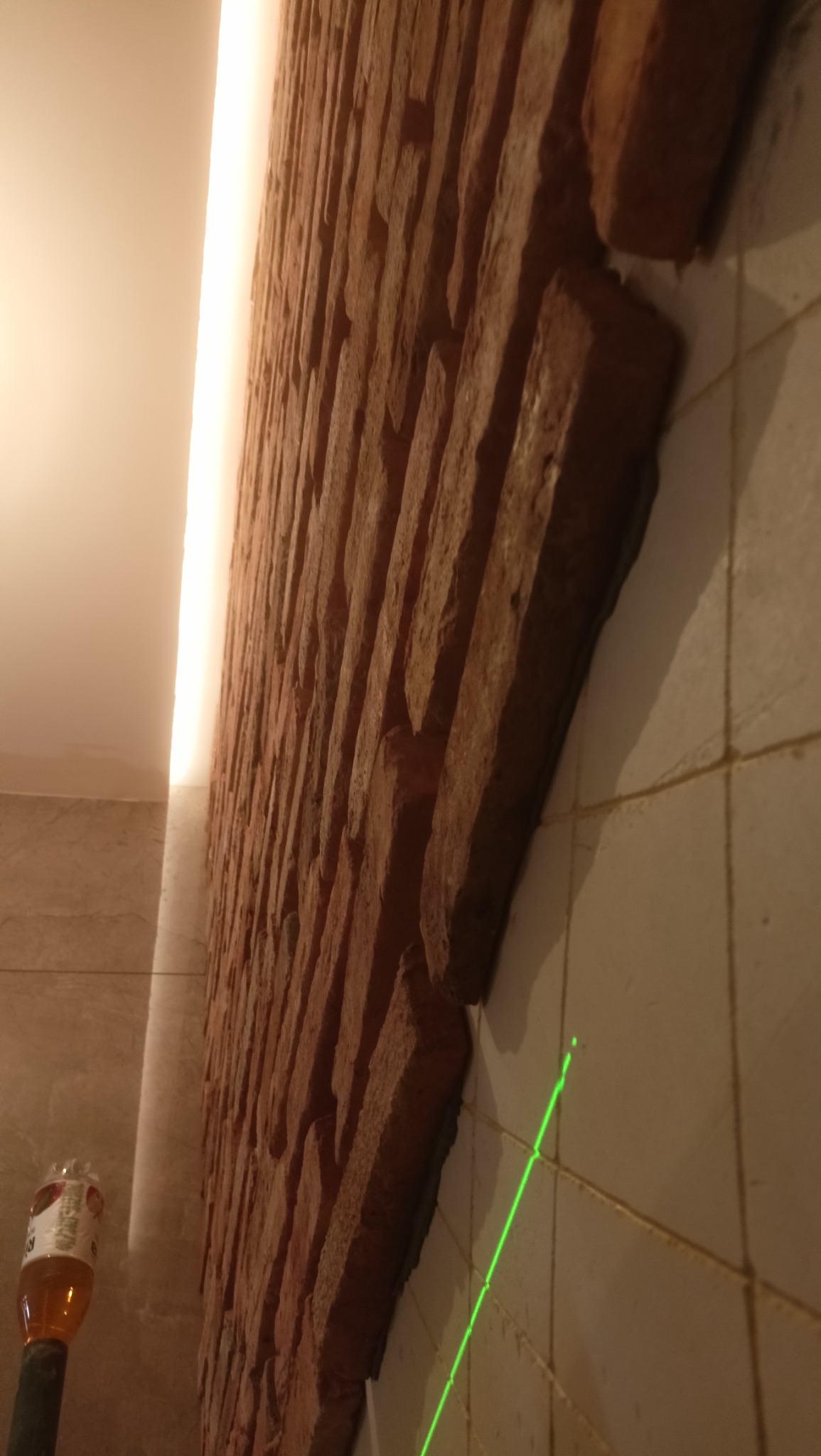 Ściana z cegły klinkierowej w trakcie montażu, widoczna wiązka lasera wyznaczająca linię, butelka z wodą widoczna w rogu.