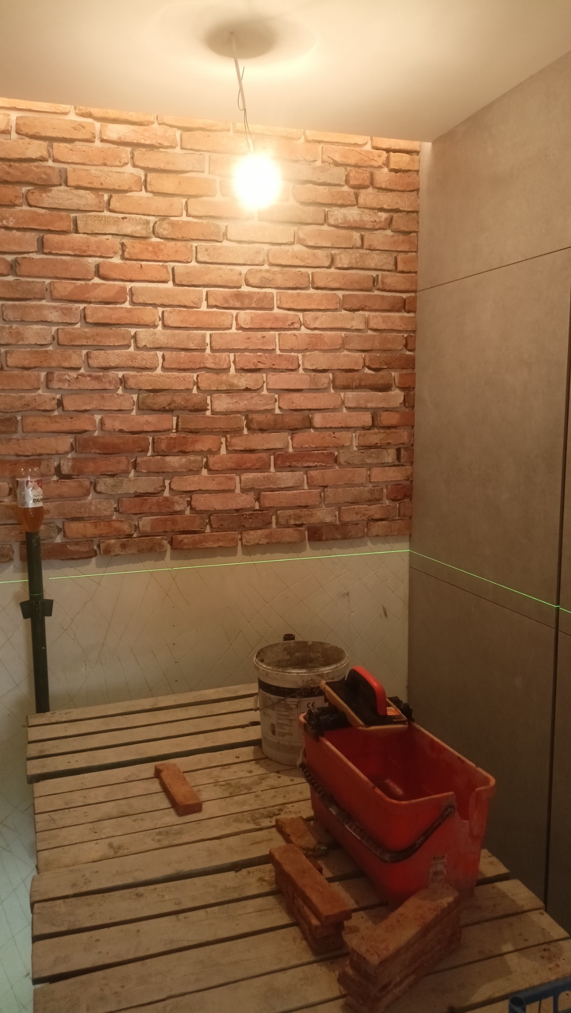 Wnętrze w trakcie wykańczania z ceglaną ścianą, widoczny laser krzyżowy, paleta robocza, wiadro i pojemnik na zaprawę, cegły przygotowane do montażu.