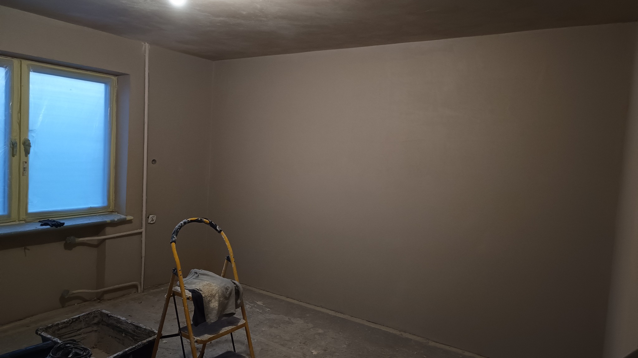 Pokój w trakcie remontu: ściany po nałożeniu gładzi, okno zabezpieczone folią, drabina z zabrudzoną szmatą, widoczne ślady po pracach na podłodze.