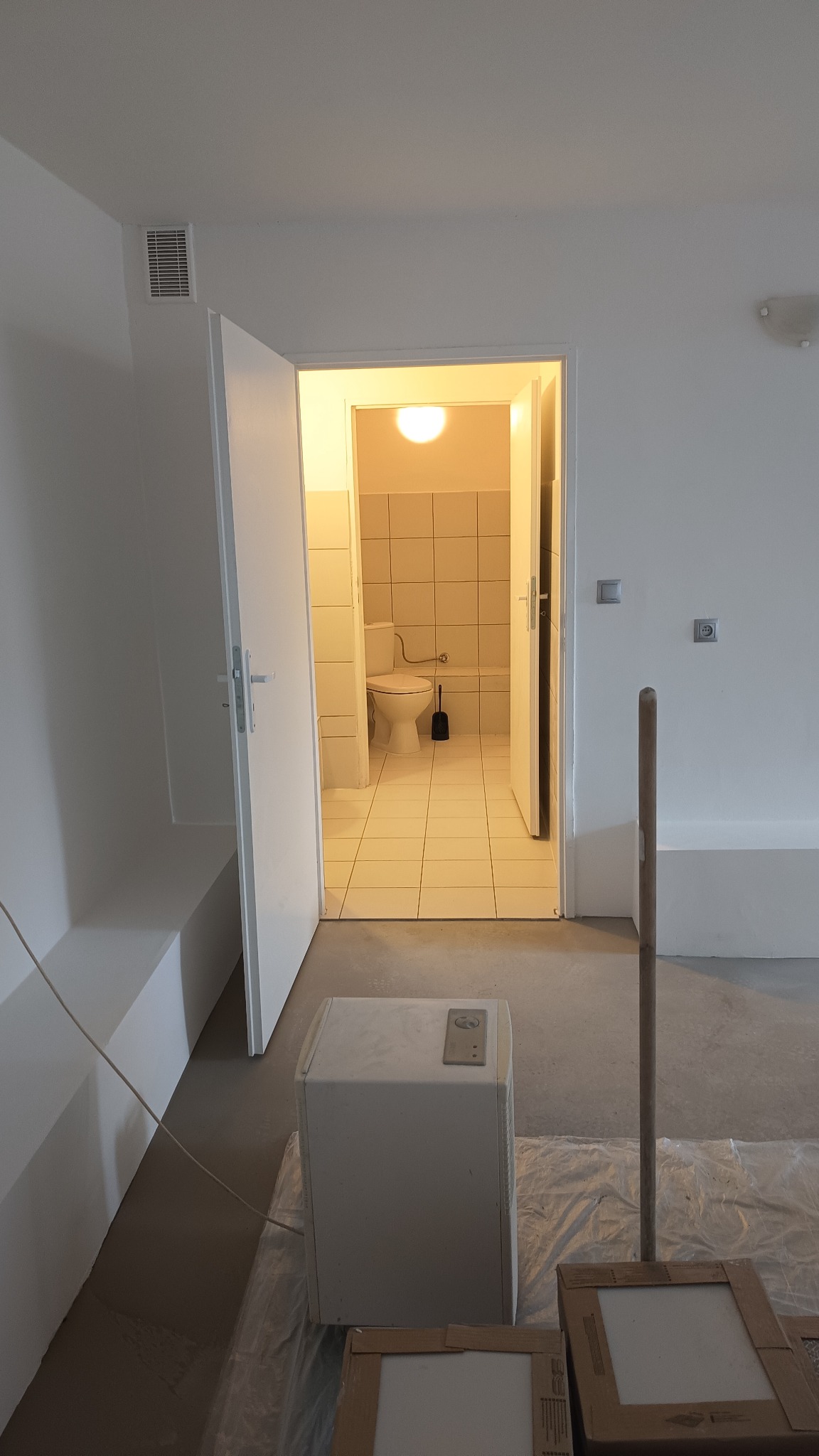 Wnętrze w trakcie prac wykończeniowych: widok na toaletę przez otwarte drzwi, podłoga zabezpieczona folią malarską, widoczny grzejnik i materiały budowlane.