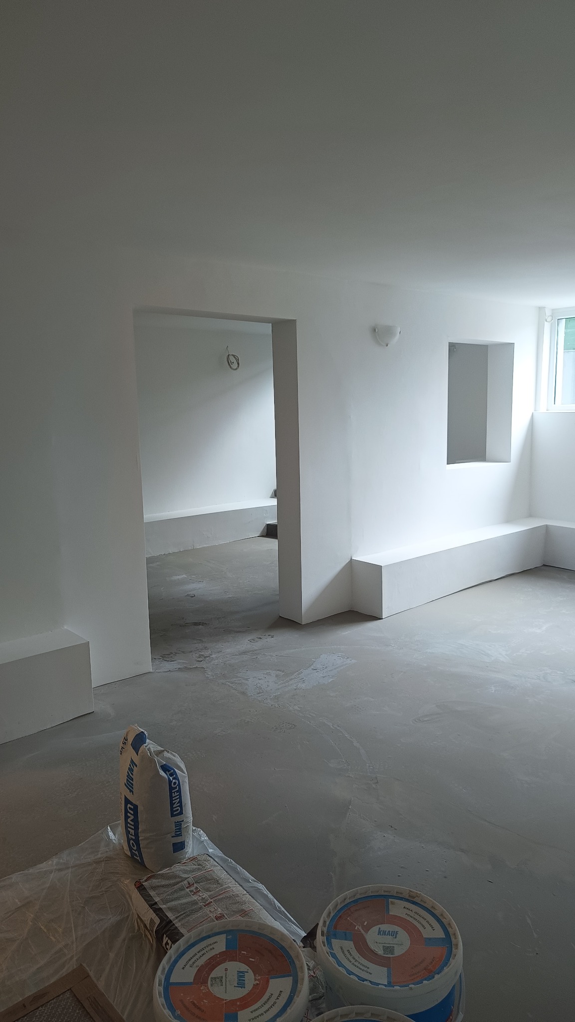 Wnętrze w trakcie remontu z białymi ścianami, widoczne worki z gładzią szpachlową Knauf Uniflott i wiadra z farbą na podłodze, surowy betonowy podkład, w tle przejście do kolejnego pomieszczenia...