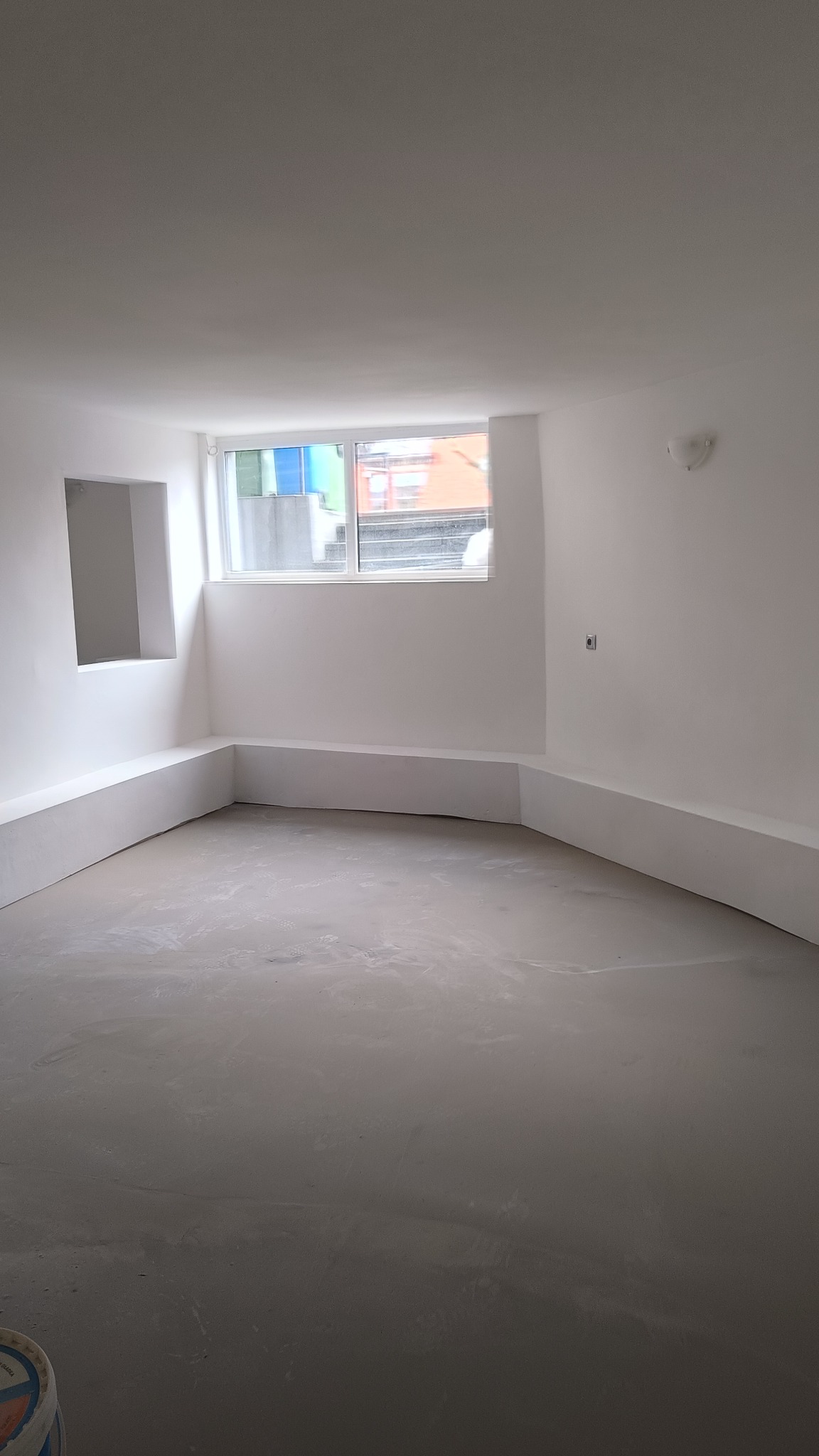 Surowe, jasne wnętrze z oknem, świeżo pomalowane na biało, z szarą, niewykończoną podłogą i wnęką w ścianie. Widoczny fragment wiadra w rogu.