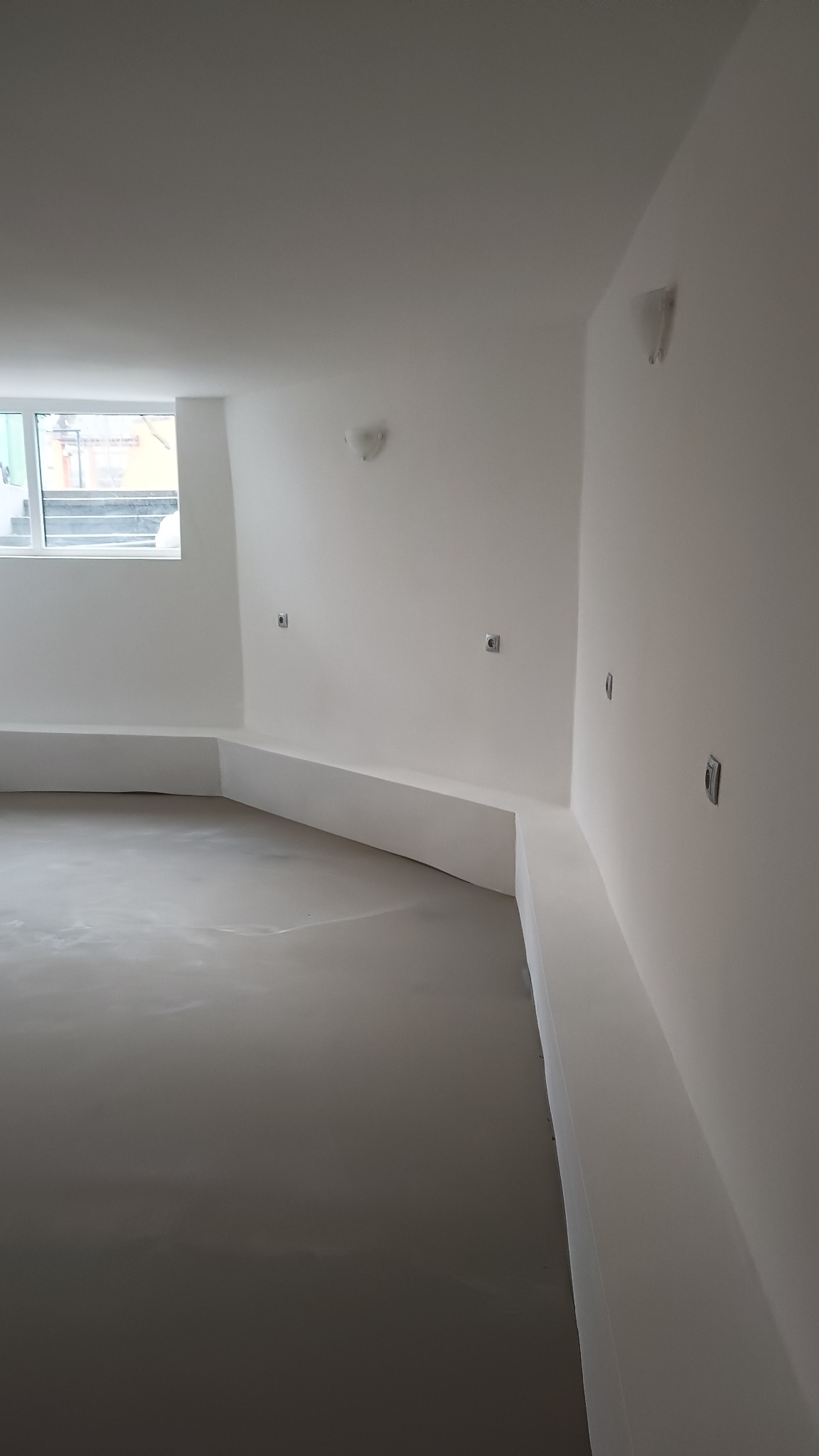 Wnętrze pokoju w Bełchatowie po wylaniu posadzki samopoziomującej, ściany przygotowane pod dalsze prace wykończeniowe, widoczne gniazdka elektryczne.
