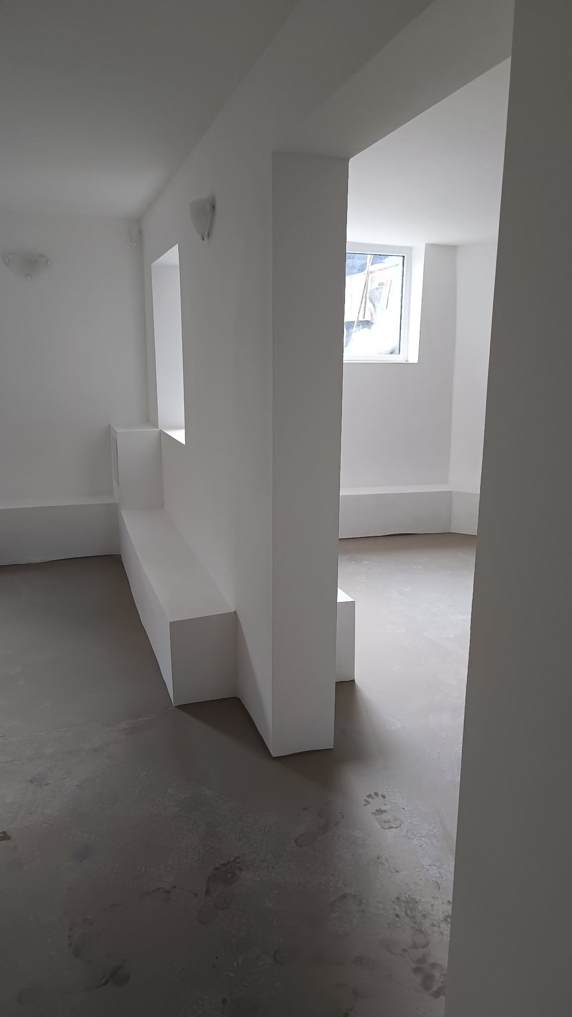 Wnętrze po świeżym pomalowaniu na biało, widoczne ślady stóp na szarej podłodze, dzieląca przestrzeń konstrukcja z półkami i oknem w tle.