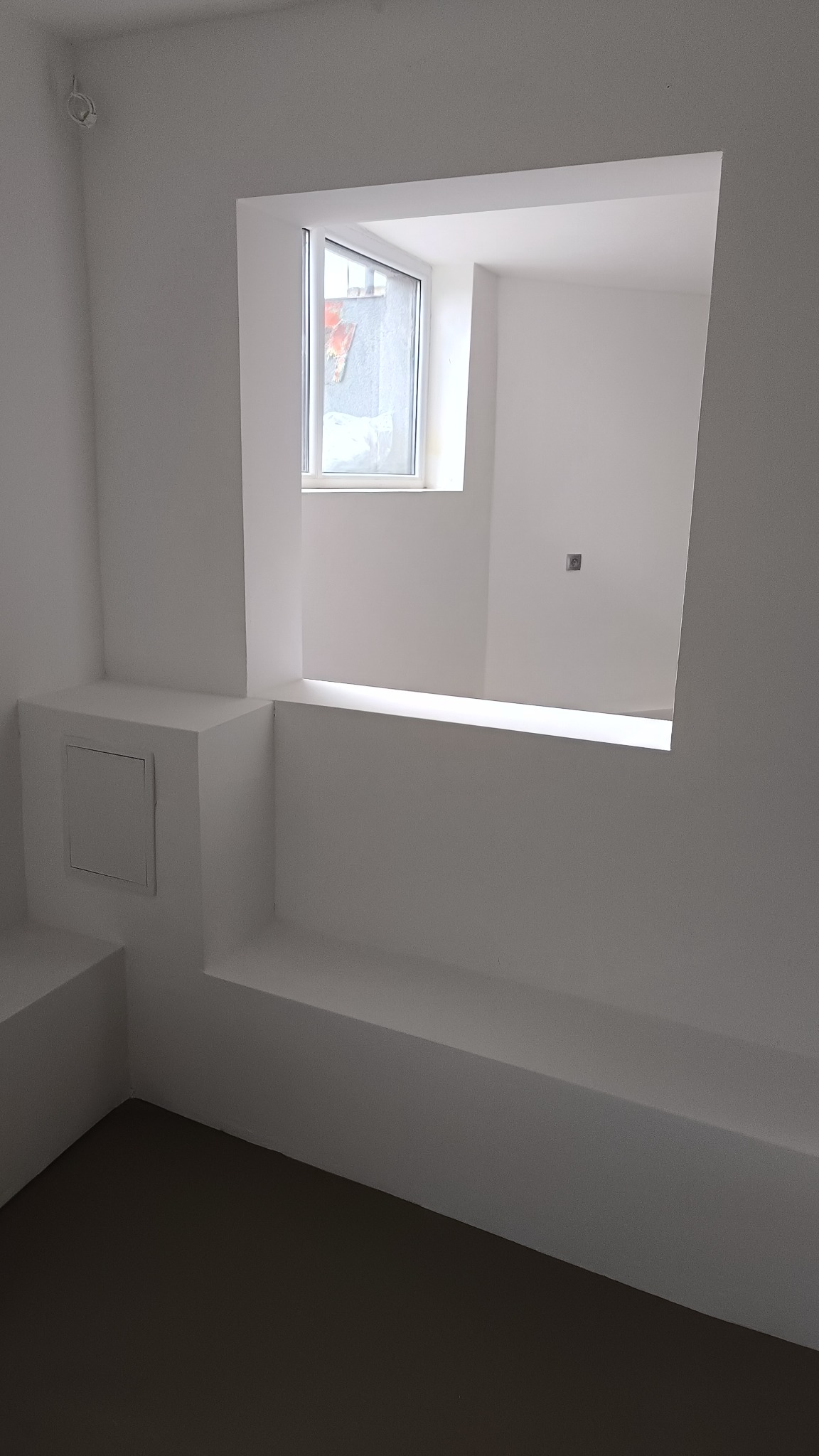 Wnętrze pomalowane na biało z oknem, widoczny schowek w ścianie z drzwiczkami oraz szary fragment podłogi.