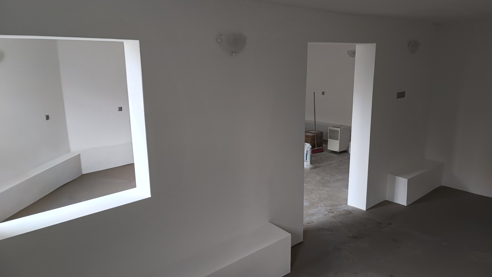 Wnętrze w trakcie remontu: świeżo pomalowane na biało ściany, widoczne otwory okienne i drzwiowe, surowa podłoga, widoczne materiały budowlane w tle.