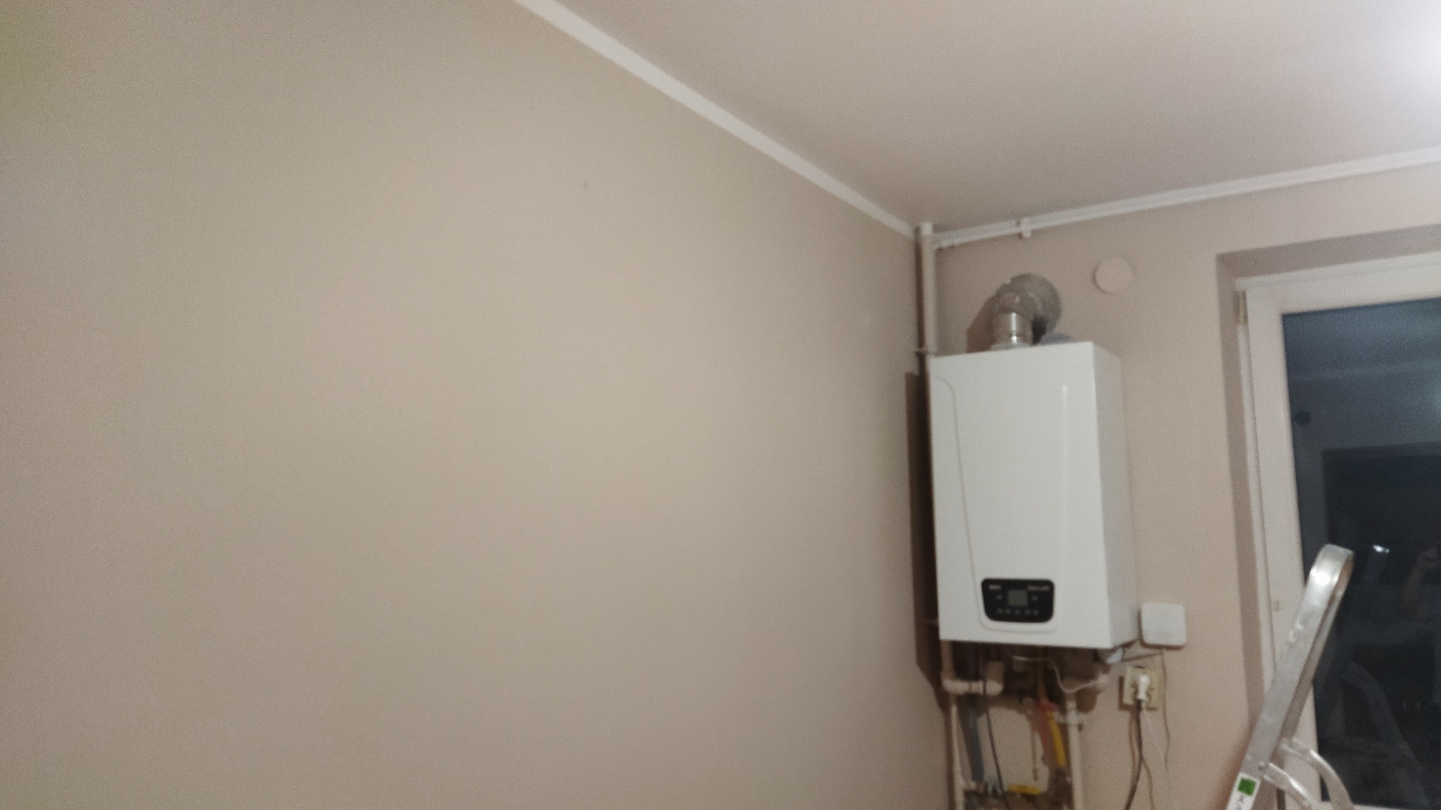 Zamontowany biały piec gazowy dwufunkcyjny na beżowej ścianie, widoczne rury, drabina aluminiowa oparta o ścianę obok okna.