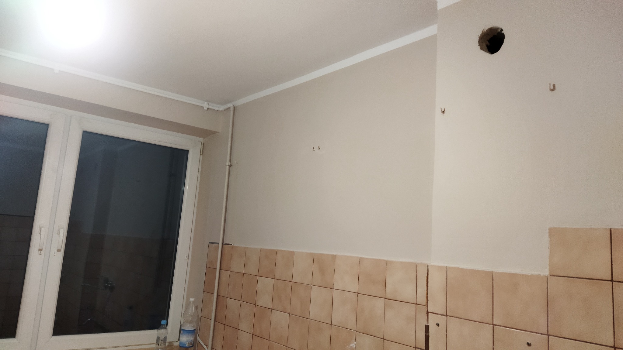 Ściana w kuchni w trakcie prac wykończeniowych: widoczne stare płytki częściowo usunięte, ściana częściowo pomalowana na beżowo, niezamaskowany otwór w ścianie, listwa sufitowa i okno z butelkami...