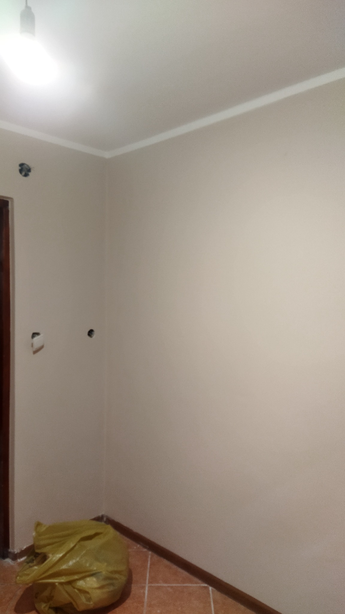 Świeżo pomalowane ściany w odcieniach beżu z widoczną listwą przysufitową i fragmentem brązowej futryny drzwi, na podłodze leży żółty worek na śmieci, widoczne otwory po gniazdkach elektrycznych.