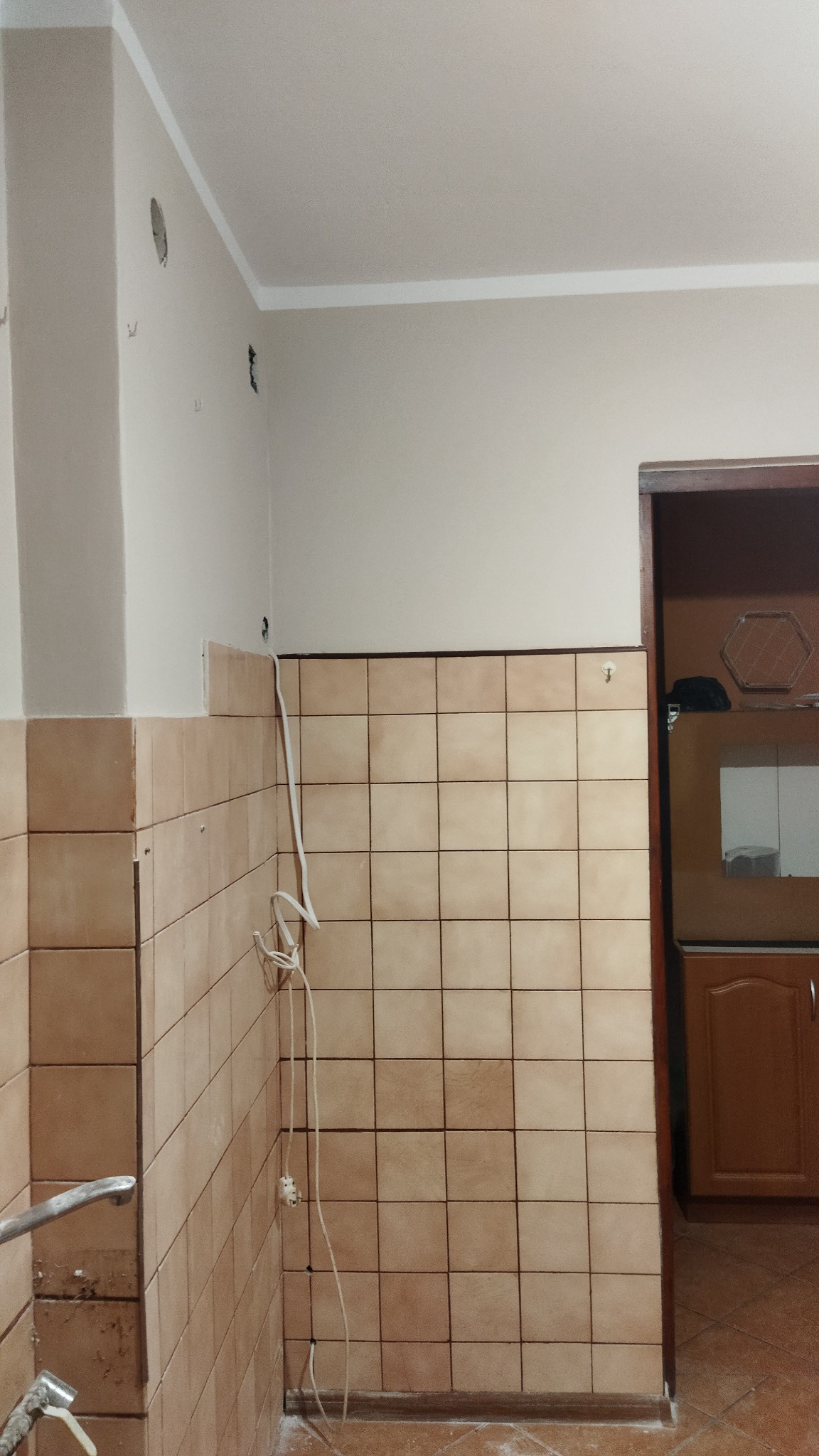 Kuchnia w trakcie remontu: beżowe płytki częściowo ułożone na ścianie, widoczne przewody elektryczne i otwory w ścianie, przygotowania do dalszych prac wykończeniowych.