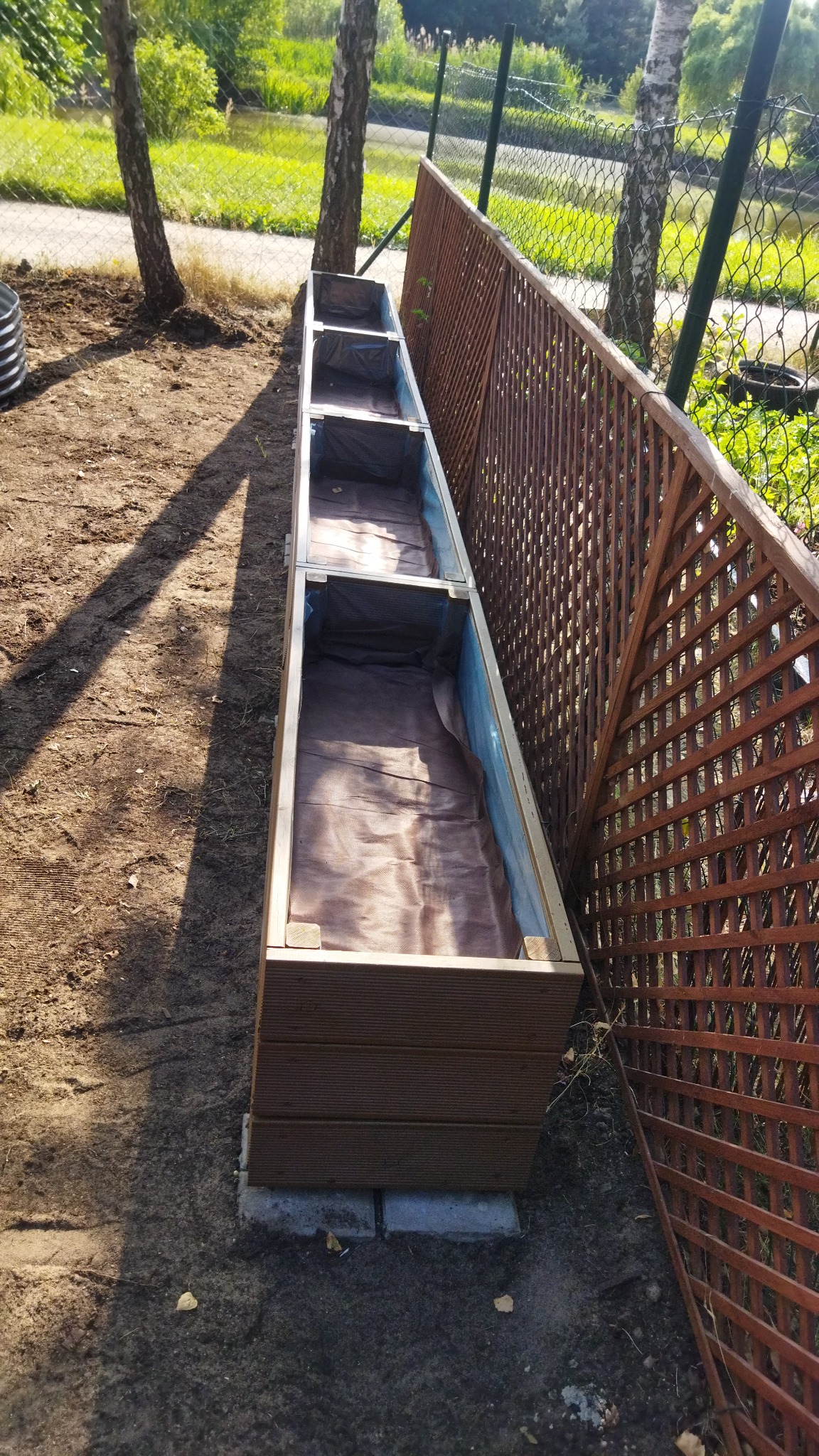 Długa, drewniana donica modułowa z brązową wyściółką, ustawiona w ogrodzie przy brązowym, ażurowym płocie, widok z góry.