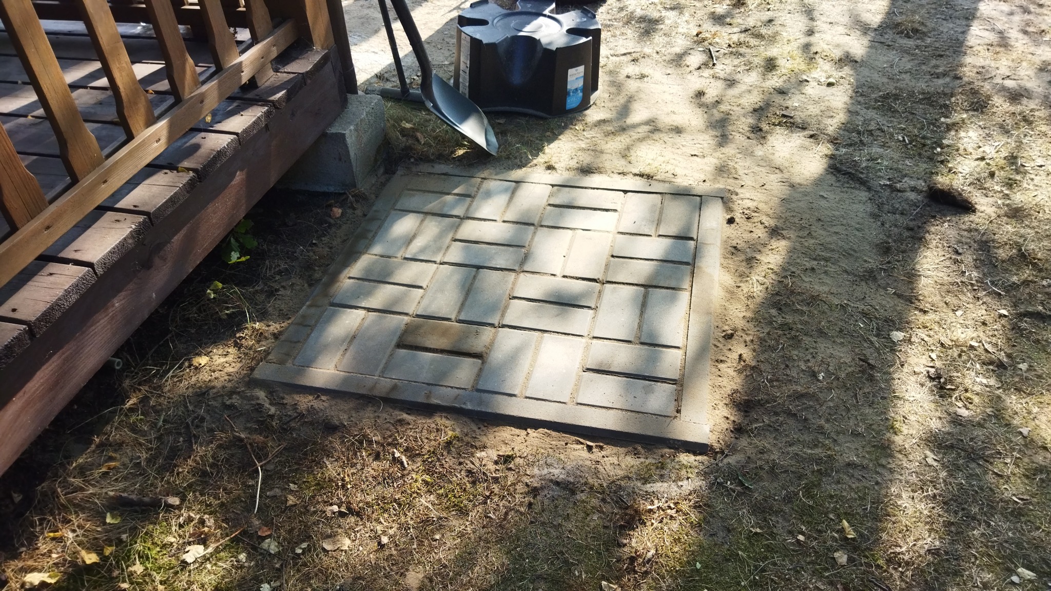 Pokrywa studzienki rewizyjnej ułożona z betonowej kostki brukowej w kształcie prostokątów, obok drewniany element konstrukcyjny, czarny zbiornik na deszczówkę i łopata.