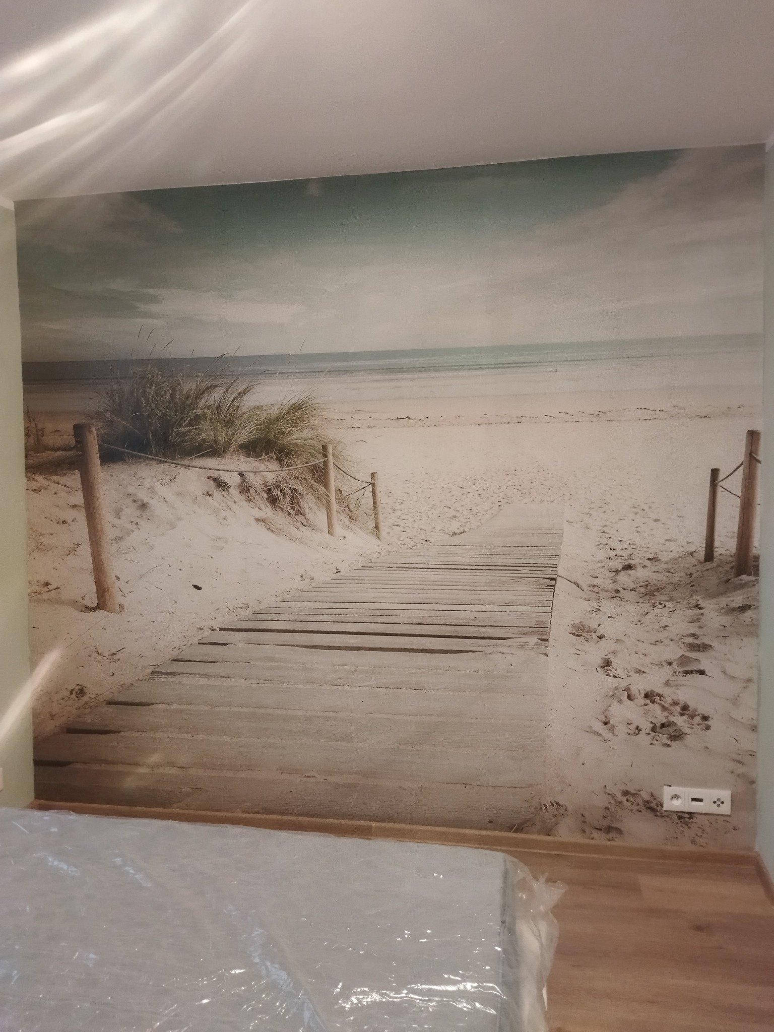 Fototapeta z motywem plaży z drewnianą ścieżką, zamontowana na ścianie w pokoju, widoczny fragment podłogi z zabezpieczeniem folią malarską oraz gniazdko elektryczne.