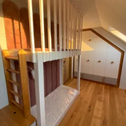 Pokój dziecięcy na poddaszu z antresolą z drabinką, zabudową szaf w skosie i drewnianą podłogą, oświetlony popołudniowym słońcem.