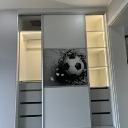 Biała szafa przesuwna z grafiką piłki nożnej, oświetleniem LED wewnątrz, z szufladami i p&oacute;łkami, na tle parkietu ułożonego w jodełkę.