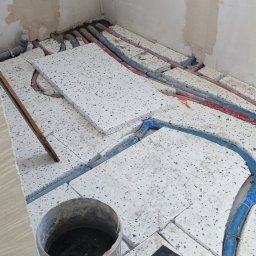 HydroMi Michał Jesionek - Montaż ogrzewania podłogowego z widocznymi rurami grzewczymi w izolacji, styropian z otworami, wiadro z wodą i folia ochronna na podłodze w surowym stanie.