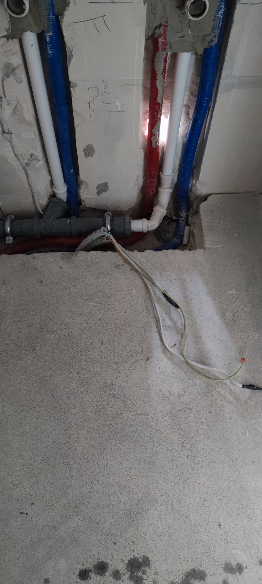 Wnęka w surowej ścianie z widocznymi rurami instalacji wodnej w kolorze białym, niebieskim i czerwonym oraz szarą rurą kanalizacyjną i przewodami elektrycznymi.