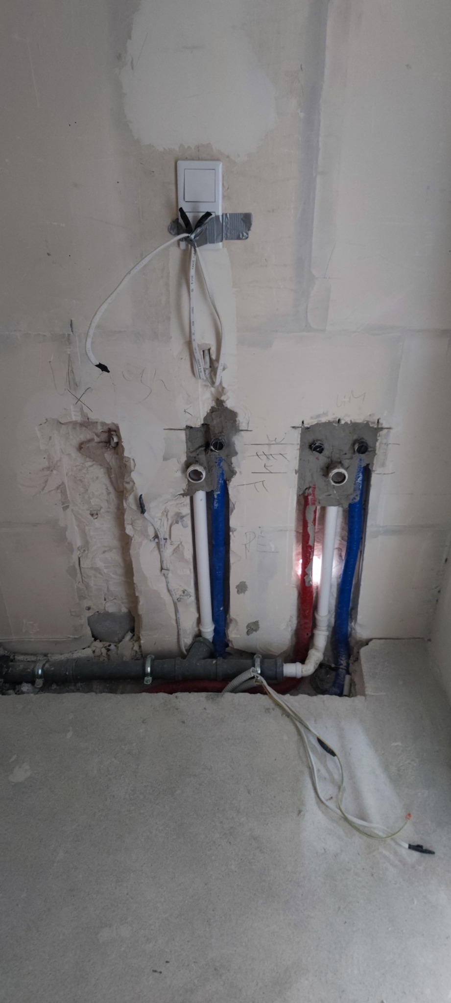 Surowy stan instalacji wodnej w ścianie: widoczne rury doprowadzające wodę ciepłą i zimną, wystające przewody elektryczne oraz nieotynkowane ściany w trakcie prac remontowych.