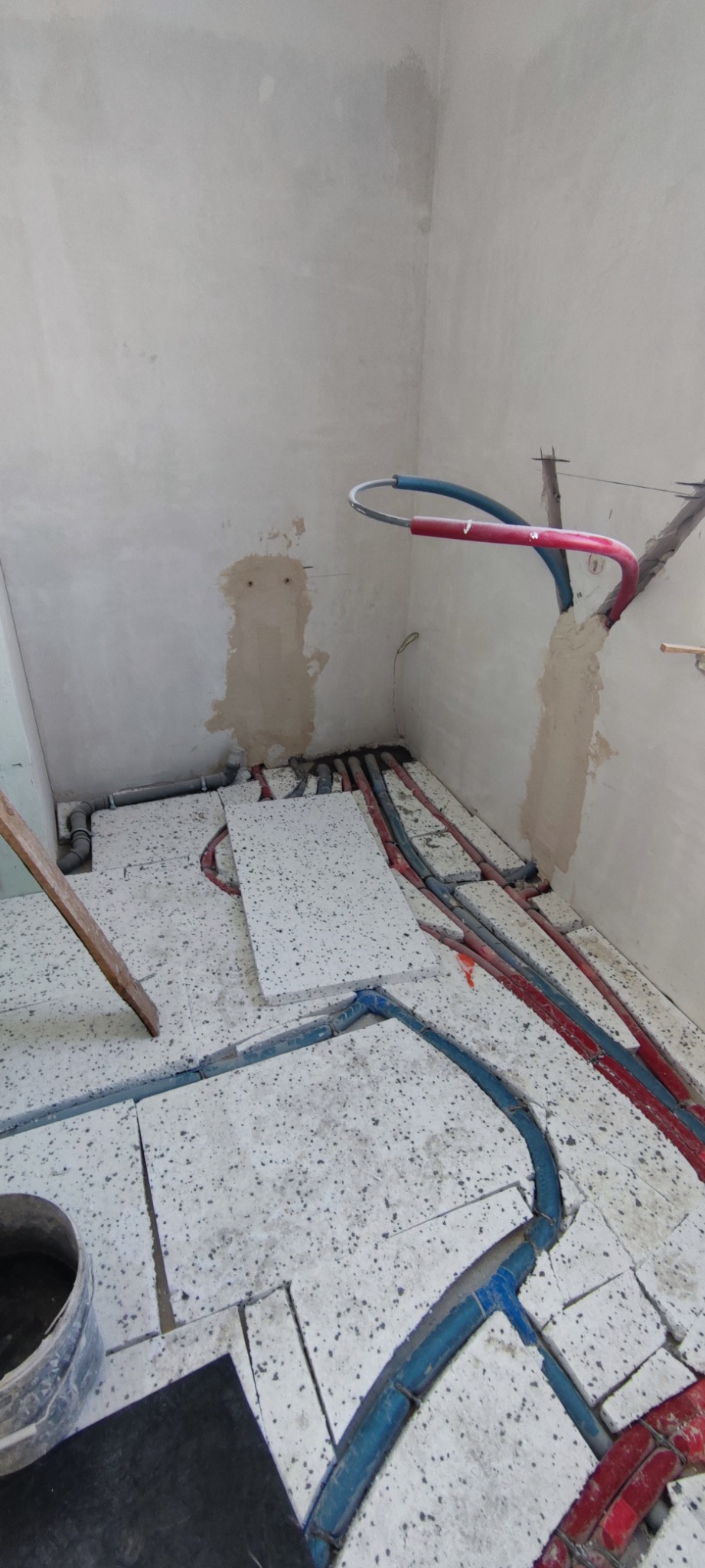 Instalacja rur grzewczych w podłodze, widoczne rury czerwone i niebieskie, częściowo zakryte płytami izolacyjnymi, ściany w stanie surowym.