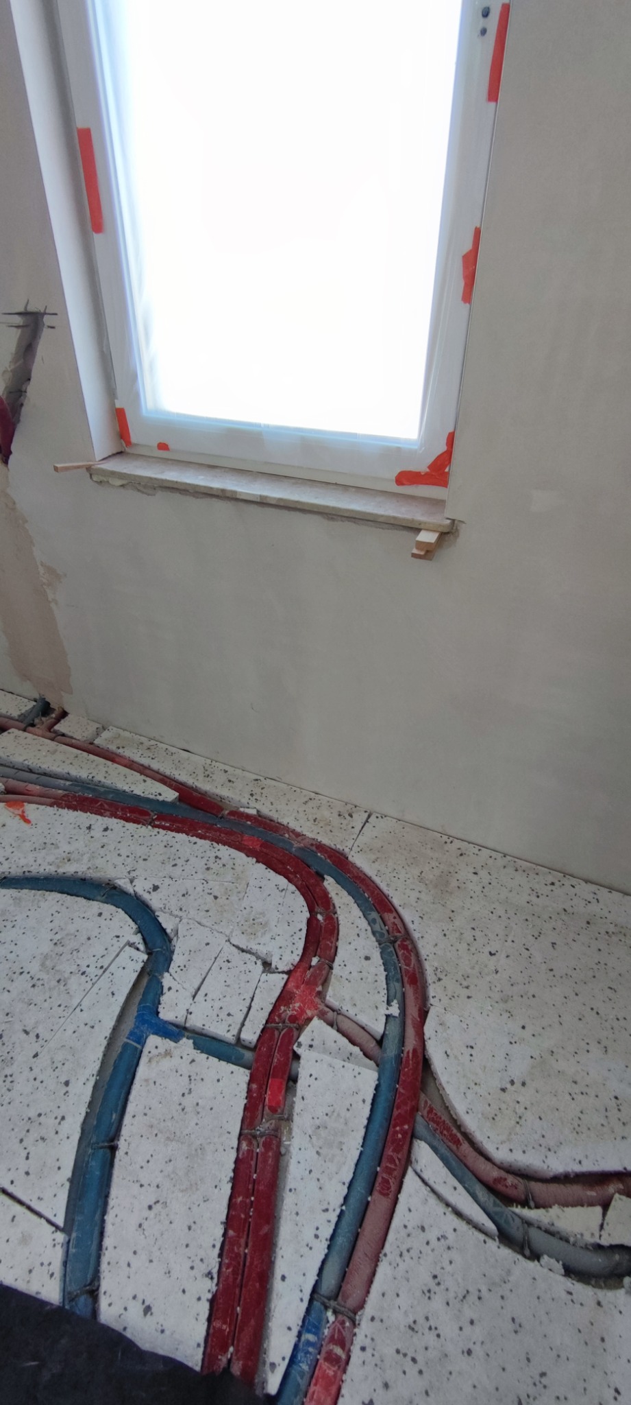 Widok na podłogę z rozłożonymi rurami ogrzewania podłogowego w kolorze czerwonym i niebieskim, przygotowanymi do zalania wylewką, w tle okno dachowe.