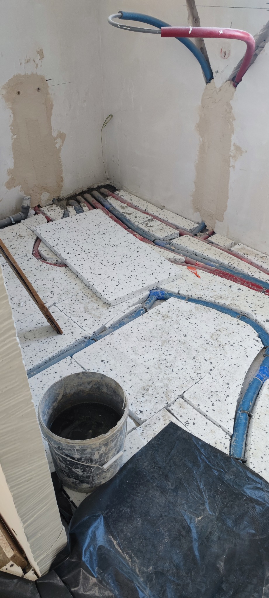 Montaż ogrzewania podłogowego z widocznymi rurami grzewczymi w izolacji, styropian z otworami, wiadro z wodą i folia ochronna na podłodze w surowym stanie.