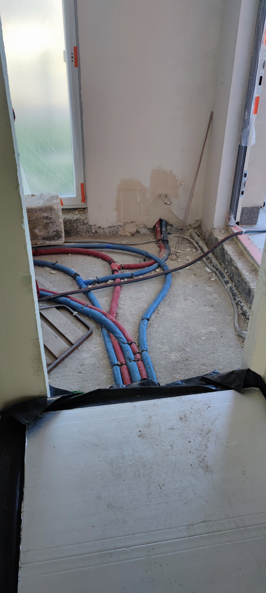 Instalacja rur grzewczych (czerwone i niebieskie) w nowym budynku, widoczne fragmenty okna i surowego wykończenia ścian.