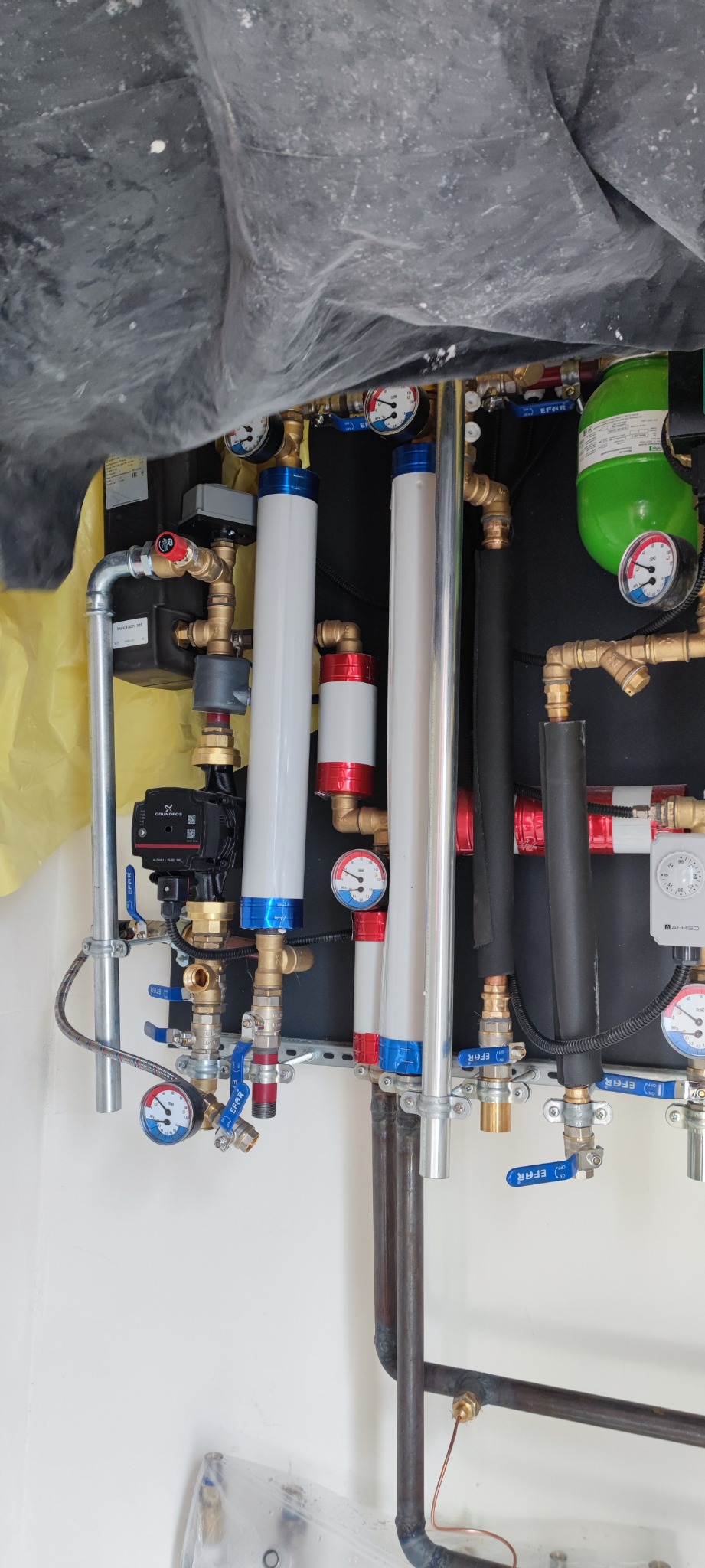 Złożona instalacja hydrauliczna z licznymi rurami miedzianymi, zaworami EFAR z niebieskimi rączkami, manometrami i pompą Grundfos, zabezpieczona folią malarską u góry.