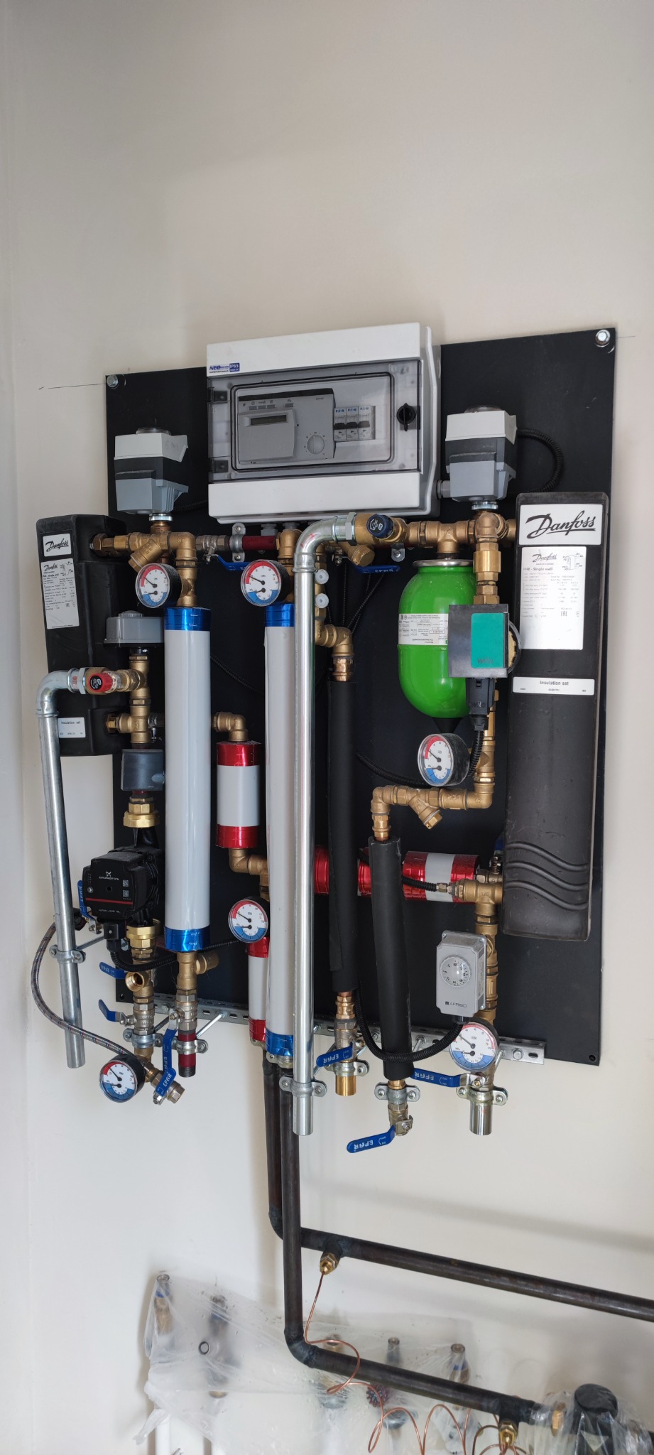 Kompletna instalacja hydrauliczna na ścianie z elementami marki Danfoss, widoczne rury miedziane, manometry, zawory, pompy obiegowe oraz sterownik elektroniczny.