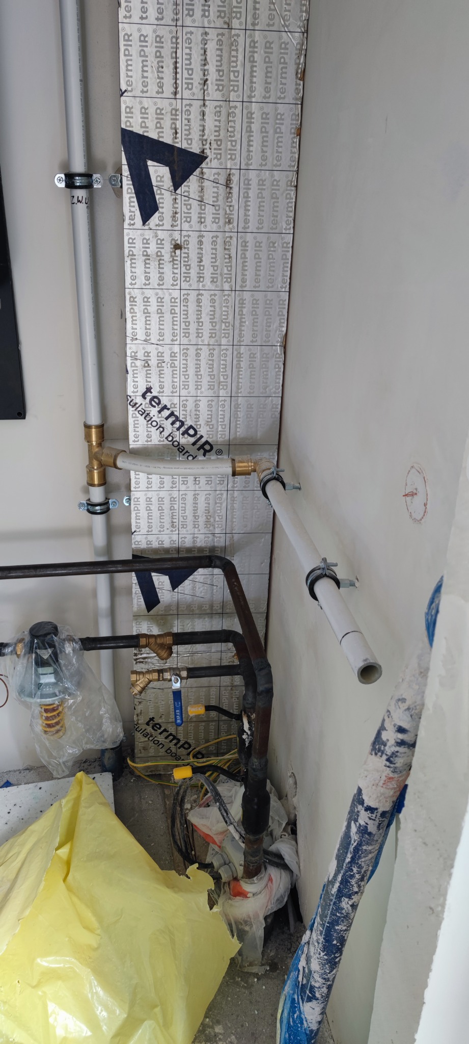 Fragment instalacji centralnego ogrzewania w trakcie montażu, widoczne rury miedziane i plastikowe, zawory, izolacja termiczna TermPIR na ścianie, oraz elementy podłączenia do grzejnika