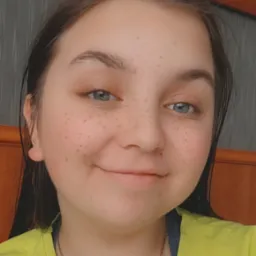 Portret uśmiechniętej nastolatki z piegami, ubrana w jaskrawożółtą koszulkę, z delikatnym naszyjnikiem na szyi, na tle drewnianego zagłówka łóżka i obrazu w ramie.