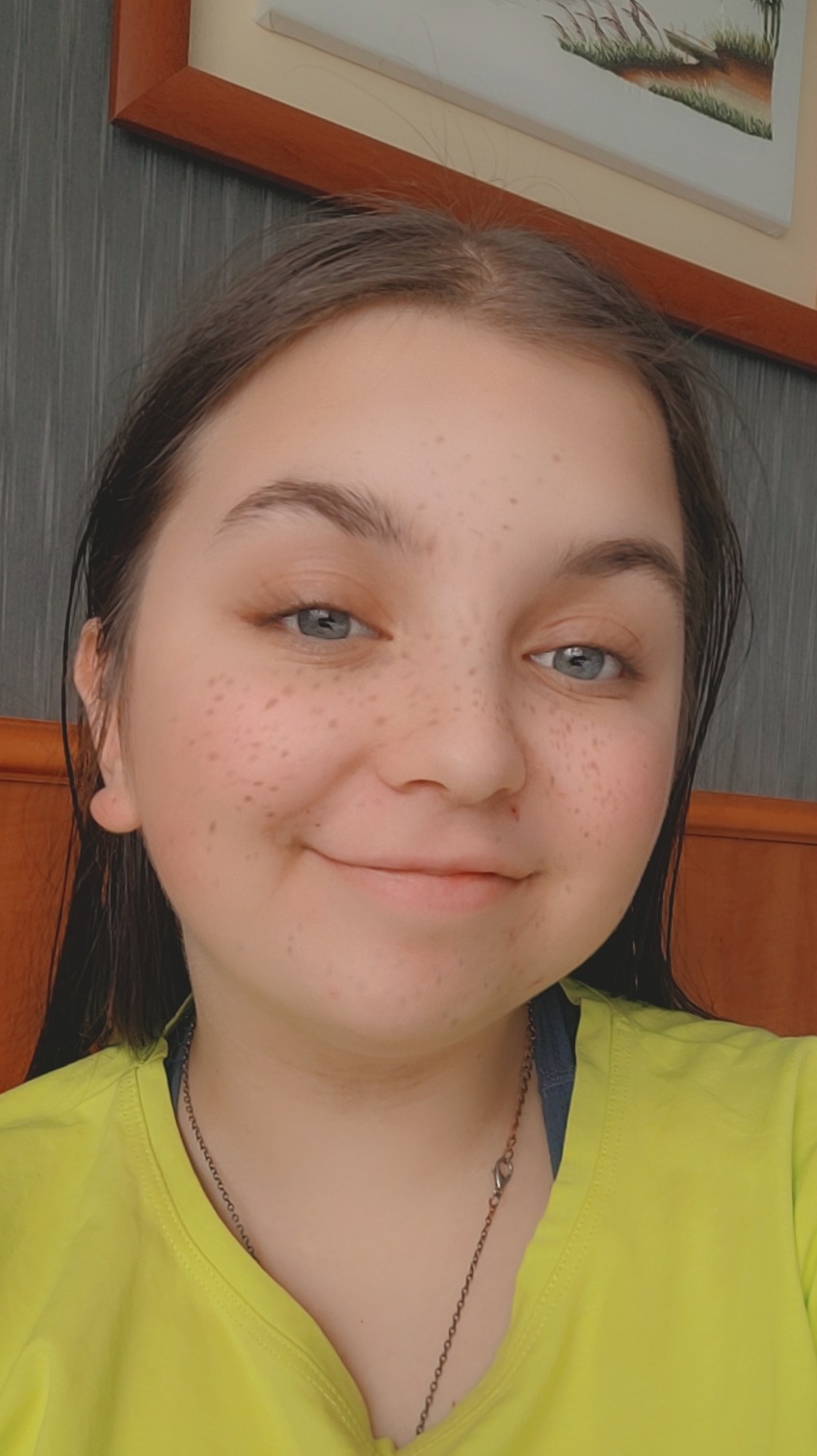 Portret uśmiechniętej nastolatki z piegami, ubrana w jaskrawożółtą koszulkę, z delikatnym naszyjnikiem na szyi, na tle drewnianego zagłówka łóżka i obrazu w ramie.