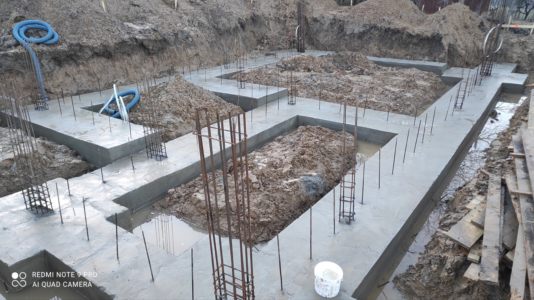 Wylane betonowe ławy fundamentowe z wystającymi prętami zbrojeniowymi, częściowo zasypane ziemią, widoczne niebieskie węże i białe wiadro na tle wykopu.