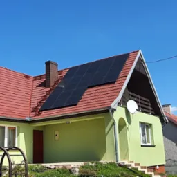 Zielony dom jednorodzinny z czerwoną dachówką i panelami słonecznymi na dachu w słoneczny dzień.