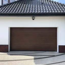 Zewnętrzny widok na segmentową bramę garażową w kolorze ciemnego drewna, zamontowaną w białym garażu w Chełmnie, z widocznymi ceglanymi detalami u podstawy ścian.