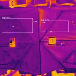 Termogram dachu budynku z zaznaczonymi obszarami i wskazaniami temperatur w stopniach Celsjusza, wykonany z lotu ptaka, widoczne samochody w tle.