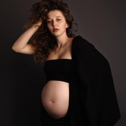 Paulina Rasińska Fotografia - Portret kobiety w ciąży, ubranej w czarny top i spodnie, z marynarką zarzuconą na ramieniu, pozującej na ciemnoszarym tle z dłonią uniesioną do kręconych włosów.
