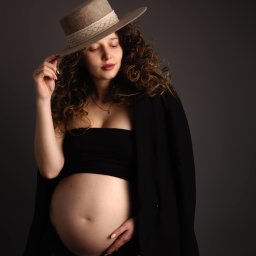 Paulina Rasińska Fotografia - Portret kobiety w ciąży, pozującej w studio w czarnym topie, marynarce i kapeluszu z szerokim rondem, dłoń trzymająca rondo kapelusza.