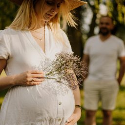Paulina Rasińska Fotografia - Kobieta w białej sukience i słomkowym kapeluszu trzyma bukiecik lawendy, dłonie spoczywają na brzuchu, w tle zamazana sylwetka mężczyzny.