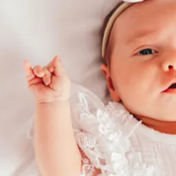 Portret noworodka w białej koronkowej sukience z kokardką na głowie, leżącego na białym tle, z uniesioną rączką.