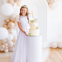 Paulina Rasińska Fotografia - Uśmiechnięta dziewczynka w białej sukience komunijnej, z wiankiem na głowie, prezentuje tort na tle dekoracji z białych i beżowych balonów.