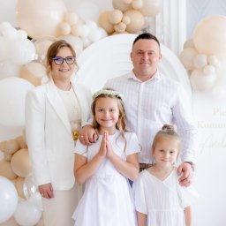 Paulina Rasińska Fotografia - Uśmiechnięta rodzina pozuje na tle dekoracji z balonów i napisu 'Pierwsza Komunia Święta'. Dziewczynka w białej sukience i wianku.