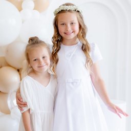 Paulina Rasińska Fotografia - Dwie uśmiechnięte dziewczynki w białych sukienkach, jedna z wiankiem, pozują na tle białych i beżowych balonów oraz białego łuku. Jasne, studyjne oświetlenie.
