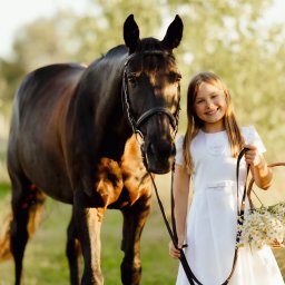 Paulina Rasińska Fotografia - Dziewczynka w białej sukience trzyma koszyk z kwiatami, stojąc obok ciemnego konia z uprzężą na tle rozmytej zieleni. Uśmiechnięta, naturalne światło.