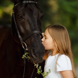 Paulina Rasińska Fotografia - Dziewczynka w białej sukience całuje czarnego konia w nos, trzymając gałązkę z zielonymi liśćmi. Ujęcie w plenerze, miękkie światło.